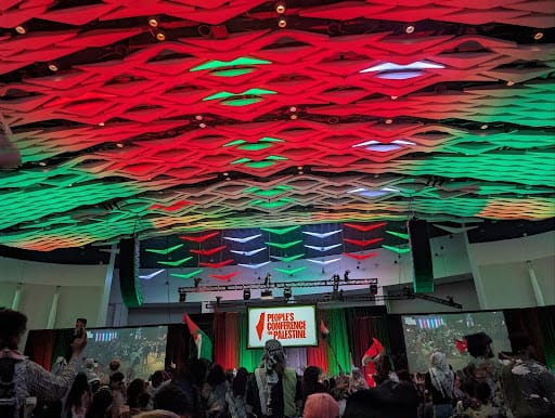 The Conference's ceiling is illuminated red, green, and white. Attendees wave Palestinian flags.